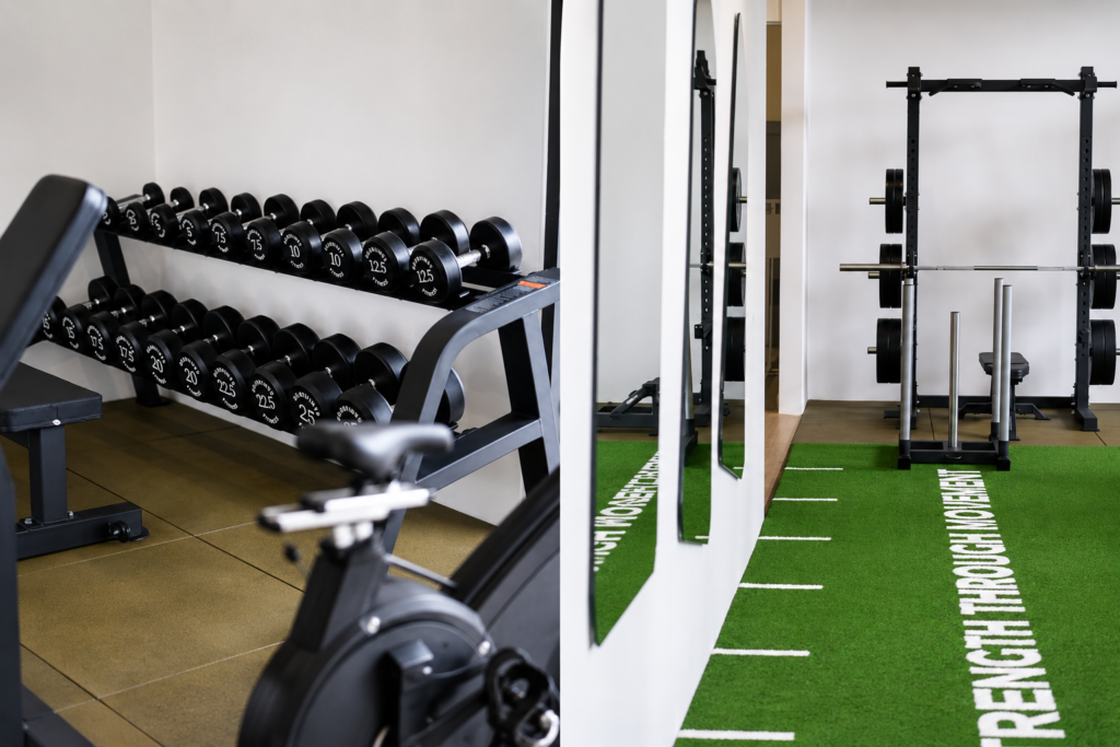 Gym with a dumbbell rack, bench, and stationary bike in foreground; green turf mat with rehabilitation text along the edge to the right.