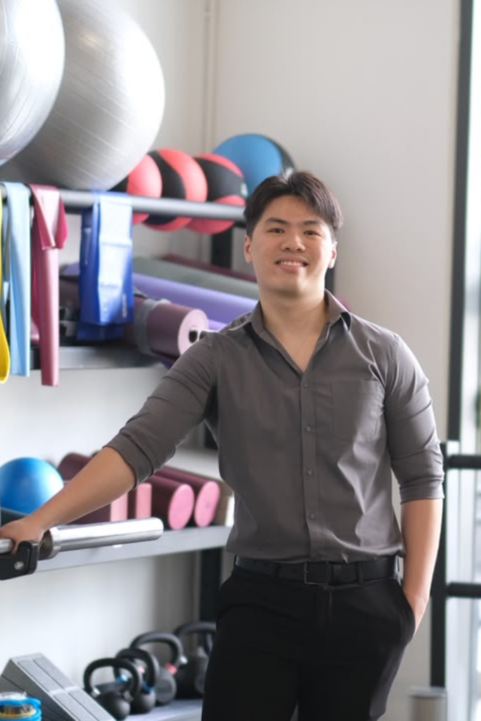 Man in a gray shirt smiling in a gym with kettlebells, mats, and workout gear on shelves behind him.